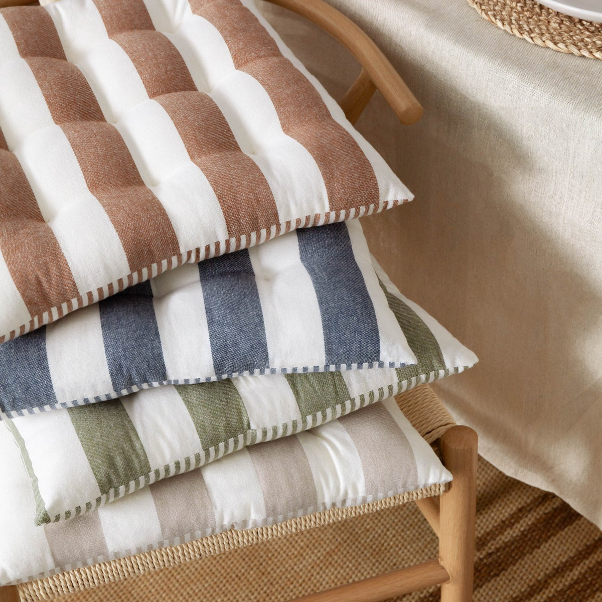 Stack of Hayle reversible cotton seat pads in rust, navy, green and beige stripes, tufted and stitched, resting on a wooden chair beside a linen tablecloth.