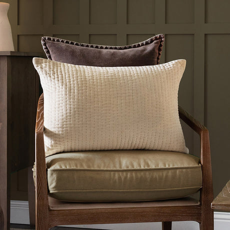 Haze Plain Velvet Cushion in Quartz, a rectangular quilted velvet cushion with ribbed texture, shown on an armchair with a brown velvet cushion behind it in a living room setting.