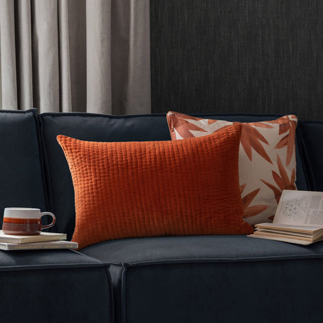 Haze Plain Velvet Cushion in Sunset orange, a rectangular quilted lumbar cushion on a navy sofa beside a leaf-print cushion, books, and a mug.