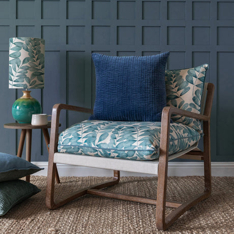 Haze Plain Velvet Cushion in Bluebelle, a quilted navy velvet square cushion styled on an armchair with leaf-patterned upholstery in a living room setting.