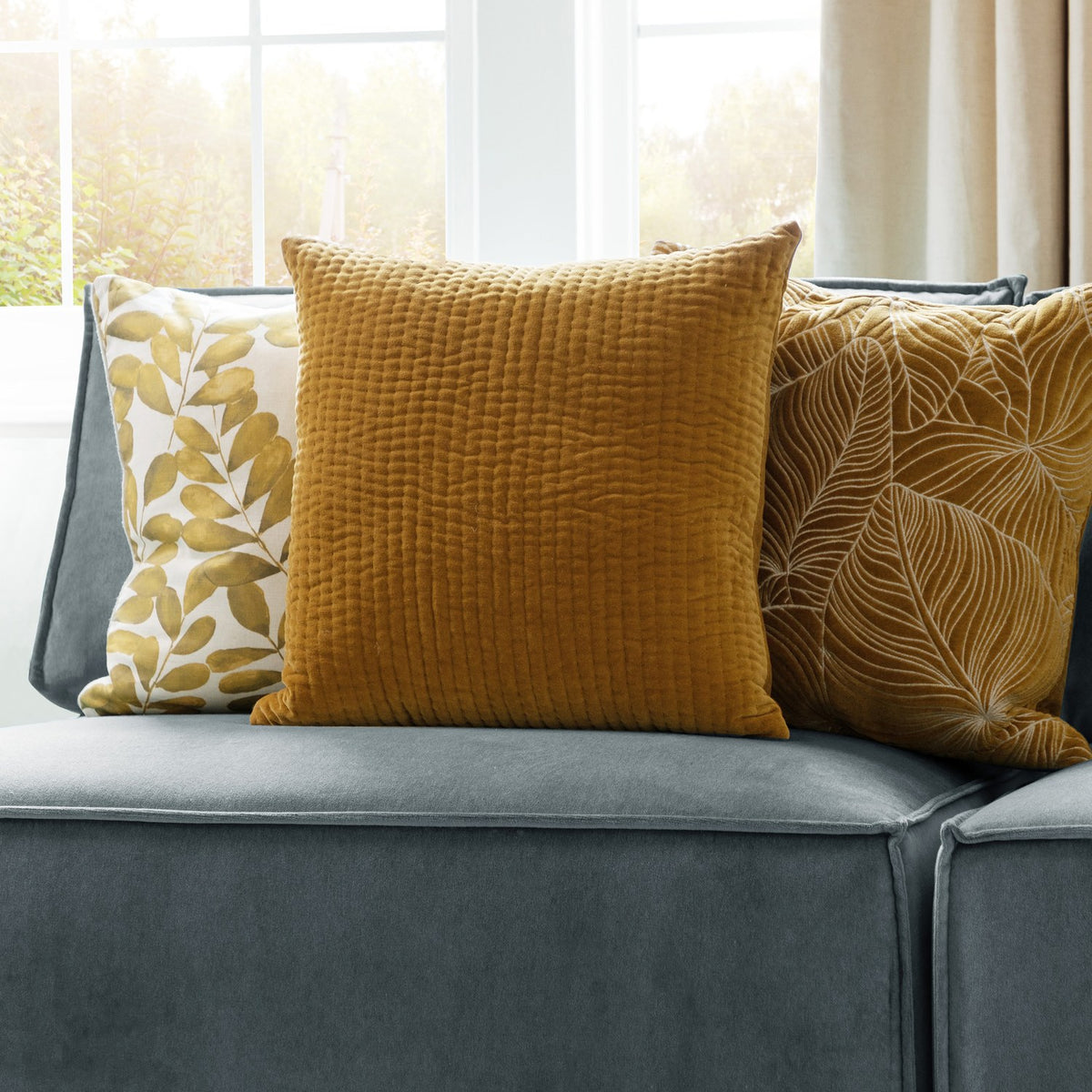 Haze Plain Velvet Cushion in mustard yellow with quilted ridge texture, shown on a grey sofa alongside patterned cushions near a bright window.