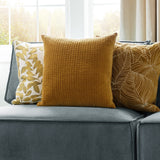 Haze Plain Velvet Cushion in mustard yellow with quilted ridge texture, shown on a grey sofa alongside patterned cushions near a bright window.