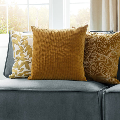 Haze Plain Velvet Cushion in mustard yellow with quilted ridge texture, shown on a grey sofa alongside patterned cushions near a bright window.