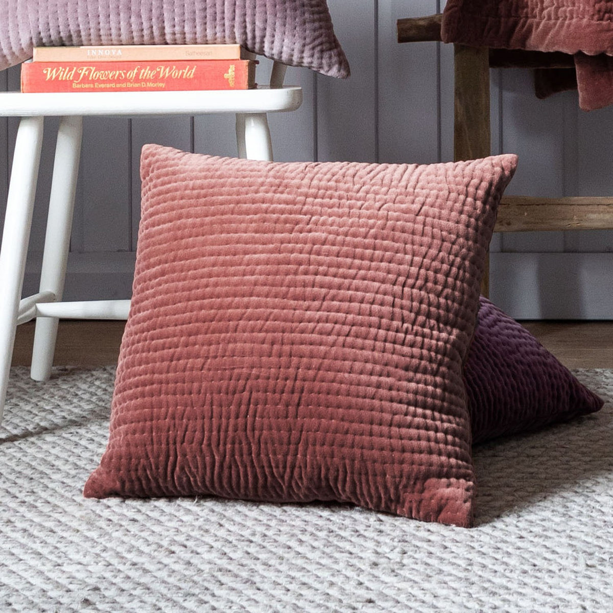 Haze Plain Velvet Cushion in Persimmon, a square quilted-front velvet cushion in warm terracotta, shown on a rug beside a stool with books in a living room setting.