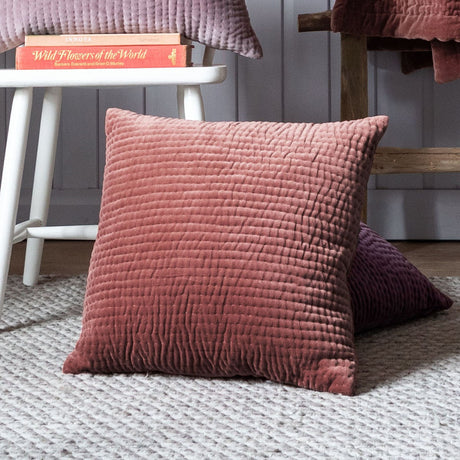 Haze Plain Velvet Cushion in Persimmon, a square quilted-front velvet cushion in warm terracotta, shown on a rug beside a stool with books in a living room setting.
