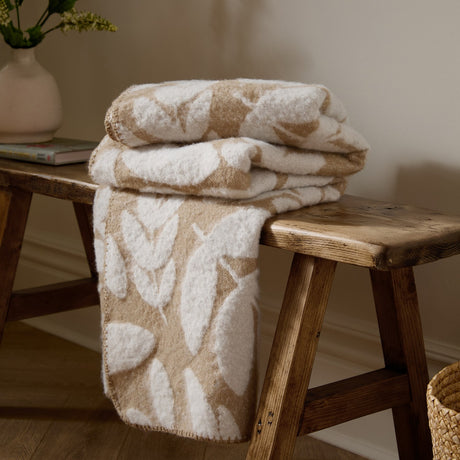Hazel Botanical Polyester Throw in oatmeal, folded and draped over a wooden bench, showing a white leaf pattern on a beige ground with blanket-stitch edging.