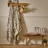 Hazel Botanical Polyester Throw in oatmeal, draped on a wooden wall-mounted coat rack with a leaf pattern and blanket-stitched edge, beside a green botanical throw in a hallway setting.