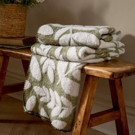 Hazel Botanical Polyester Throw in olive green with white leaf pattern, folded on a wooden stool beside books and a potted plant.