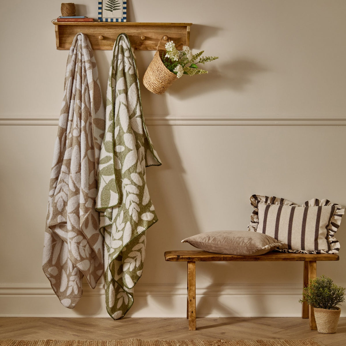 Hazel Botanical Polyester Throw in olive green with cream leaf pattern, draped on a wall-mounted hook beside a matching beige throw in a hallway setting.
