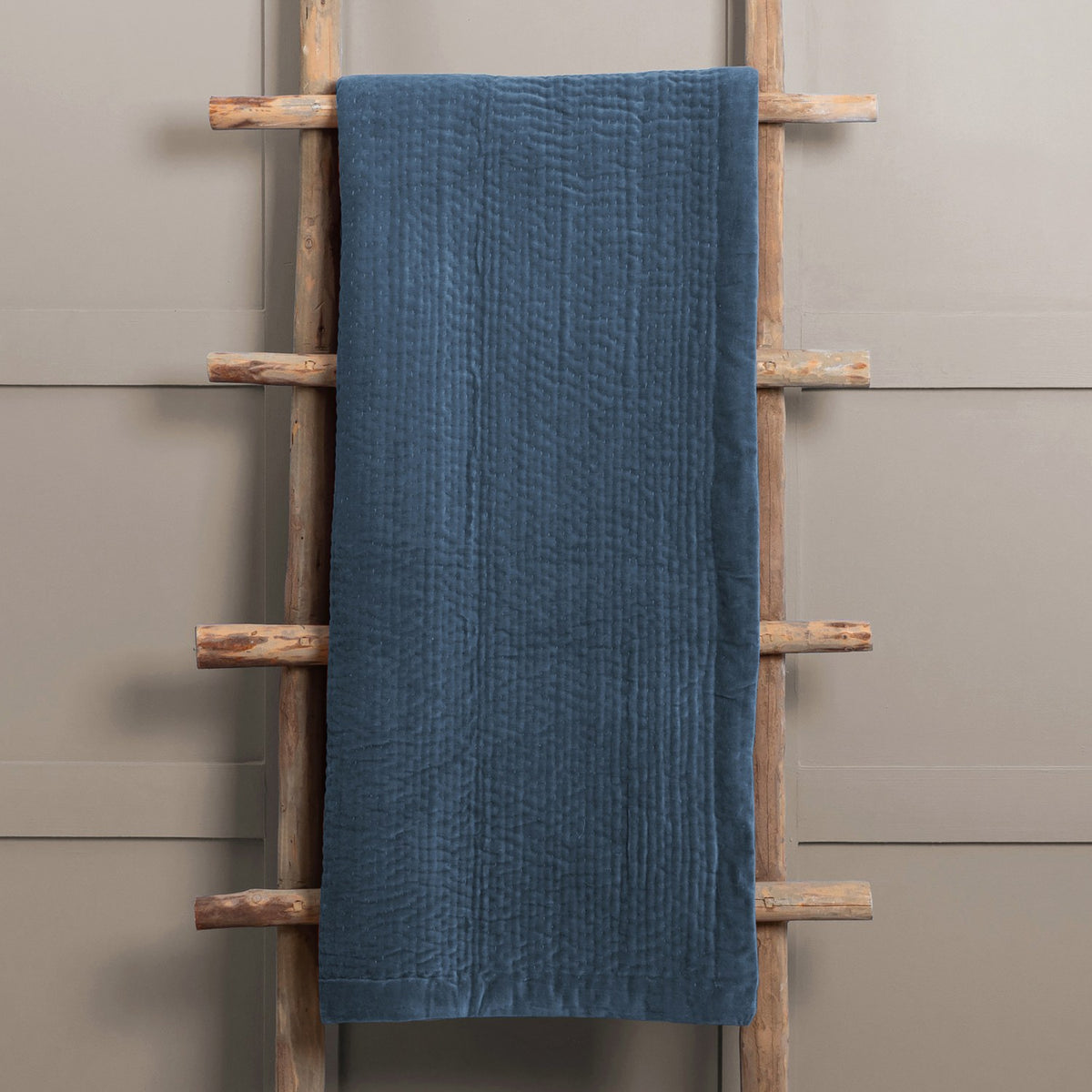 Haze Velvet Cotton Throw in Bluebell blue, quilted cotton velvet with vertical stitched texture, draped over a wooden ladder against a neutral wall.