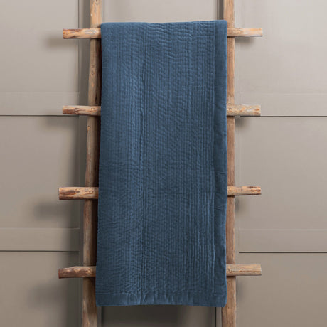 Haze Velvet Cotton Throw in Bluebell blue, quilted cotton velvet with vertical stitched texture, draped over a wooden ladder against a neutral wall.