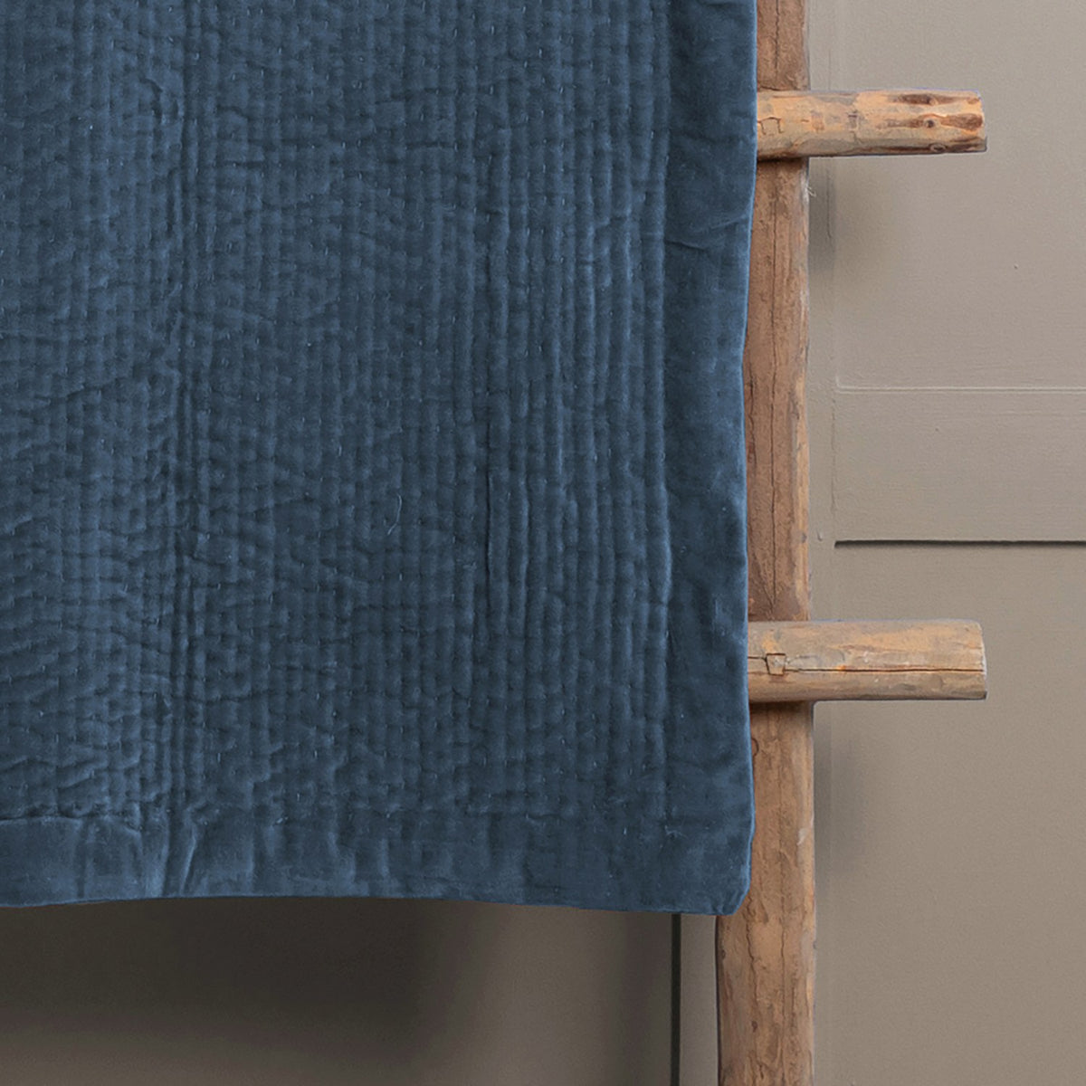 Haze Velvet Cotton Throw in bluebell blue, quilted cotton velvet with textured stitched rows, draped over a wooden ladder against a grey wall.
