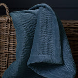 Haze Velvet Cotton Throw in bluebell blue, quilted cotton velvet with hand-stitched texture, draped over a wicker basket against a dark background.