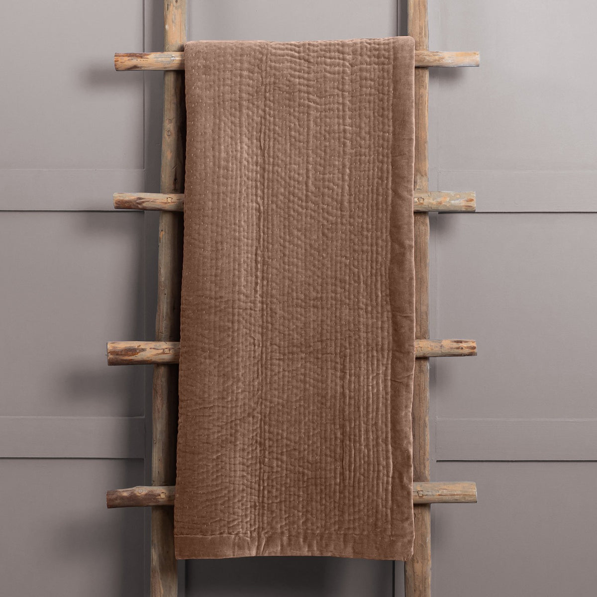 Haze Velvet Cotton Throw in brown, quilted velvet texture with visible stitched rows, draped over a wooden ladder against a grey wall.