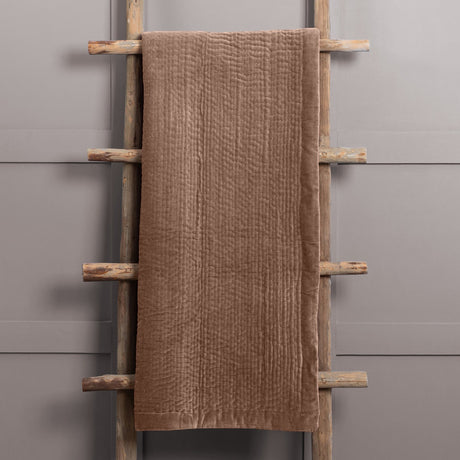 Haze Velvet Cotton Throw in brown, quilted velvet texture with visible stitched rows, draped over a wooden ladder against a grey wall.