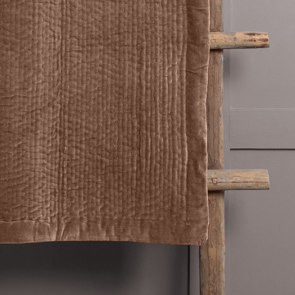 Haze Velvet Cotton Throw in brown, quilted velvet cotton with hand-stitched texture, draped over a wooden ladder against a grey wall.