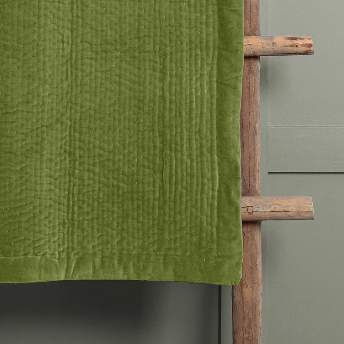 Haze Velvet Cotton Throw in grass green, hand-quilted cotton velvet with rippling texture and smooth border edge, draped over a wooden ladder against a grey wall.