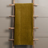 Haze Velvet Cotton Throw in mustard, hand-quilted cotton velvet with rippled texture, draped over a wooden ladder against a grey background.