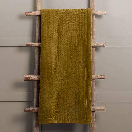 Haze Velvet Cotton Throw in mustard, hand-quilted cotton velvet with rippled texture, draped over a wooden ladder against a grey background.