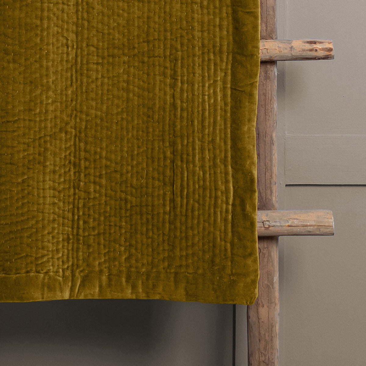 Haze Velvet Cotton Throw in mustard, hand-quilted cotton velvet with rippled texture, draped over a rustic wooden ladder against a grey wall.
