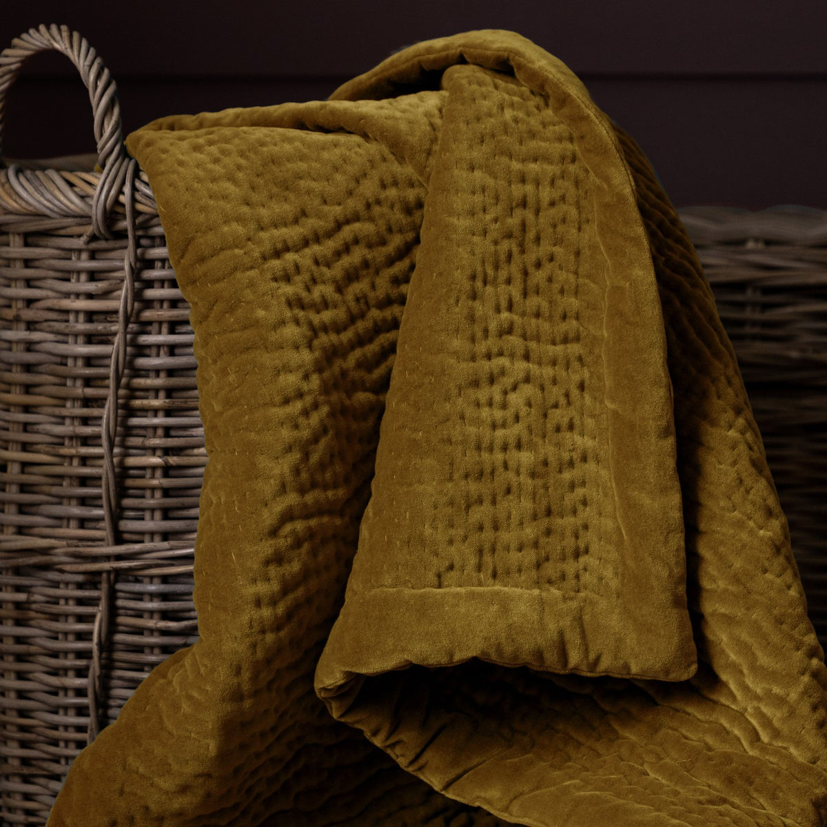 Haze Velvet Cotton Throw in mustard, hand-quilted rippled texture, draped over a woven wicker basket in a dark-toned room.