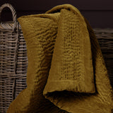 Haze Velvet Cotton Throw in mustard, hand-quilted rippled texture, draped over a woven wicker basket in a dark-toned room.