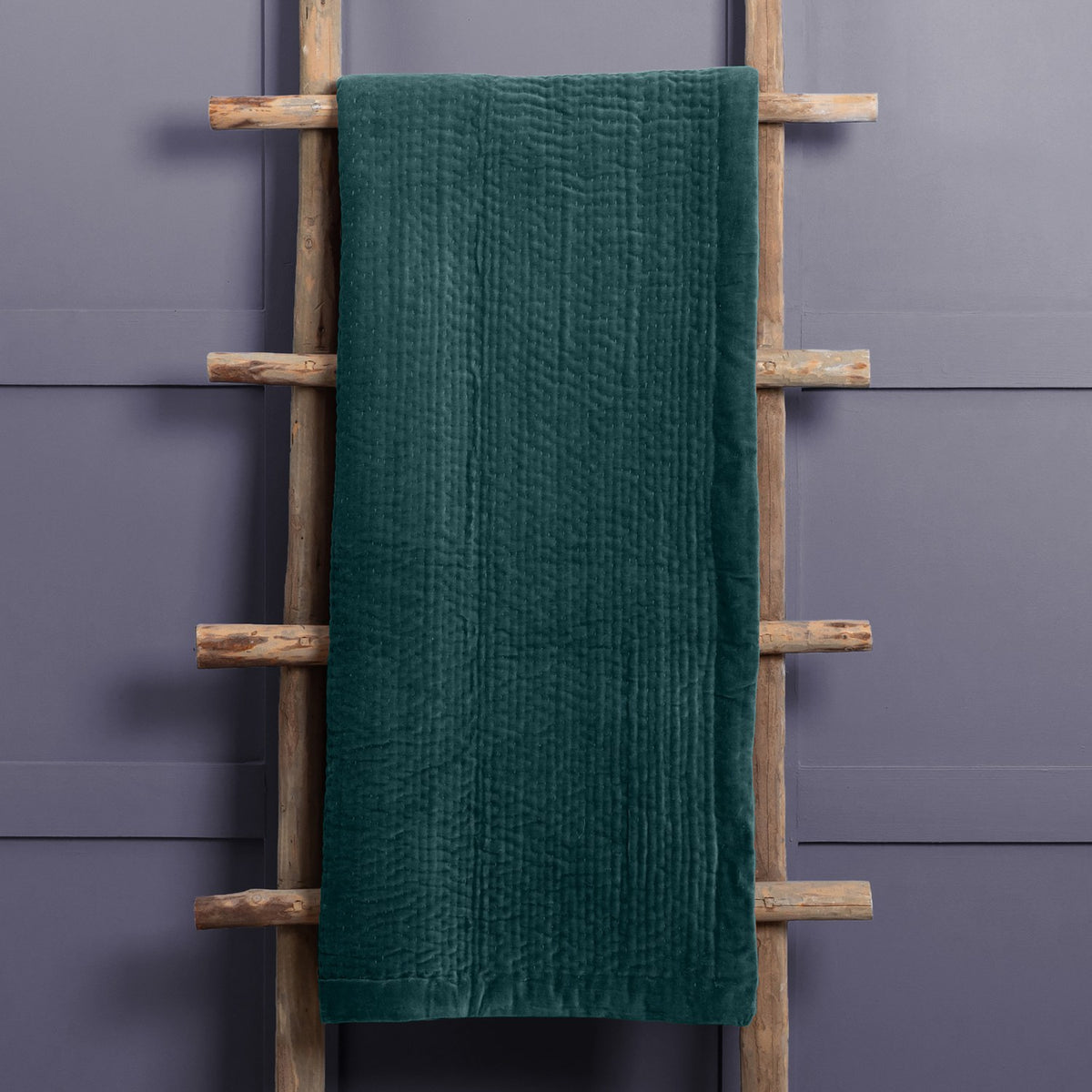 Haze Velvet Cotton Throw in Ocean, a deep teal quilted cotton throw with rippled texture, draped over a wooden ladder against a grey wall.