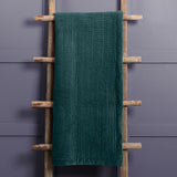 Haze Velvet Cotton Throw in Ocean, a deep teal quilted cotton throw with rippled texture, draped over a wooden ladder against a grey wall.