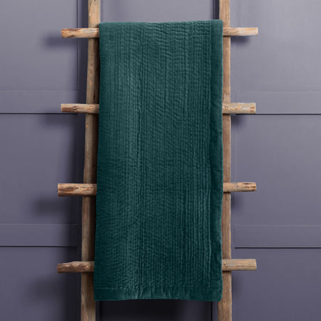 Haze Velvet Cotton Throw in Ocean, a deep teal quilted cotton throw with rippled texture, draped over a wooden ladder against a grey wall.