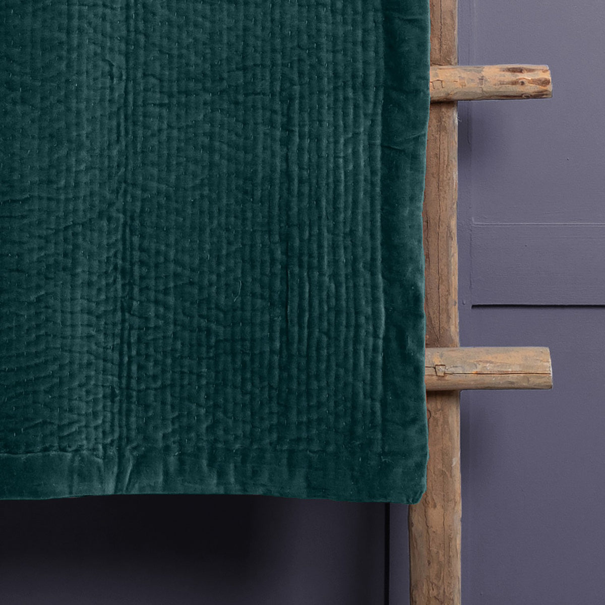 Haze Velvet Cotton Throw in Ocean, a hand-quilted teal velvet throw with rippled texture, draped over a wooden ladder against a grey wall.