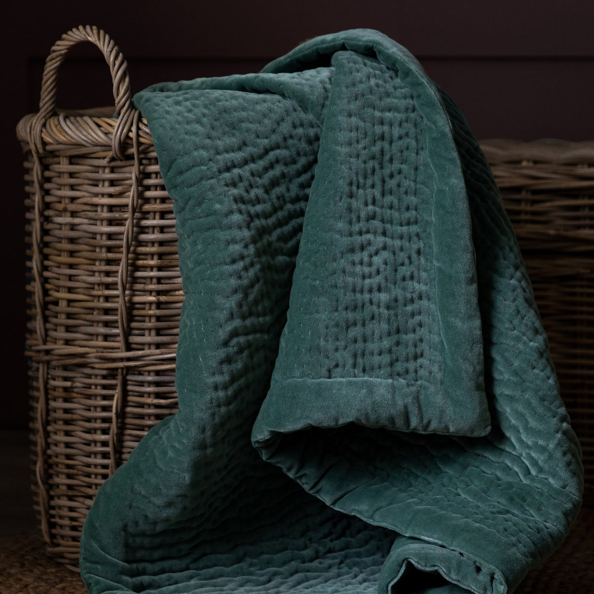 Haze Velvet Cotton Throw in Ocean, a deep teal quilted velvet throw with rippled hand-quilted texture, draped over woven wicker baskets.