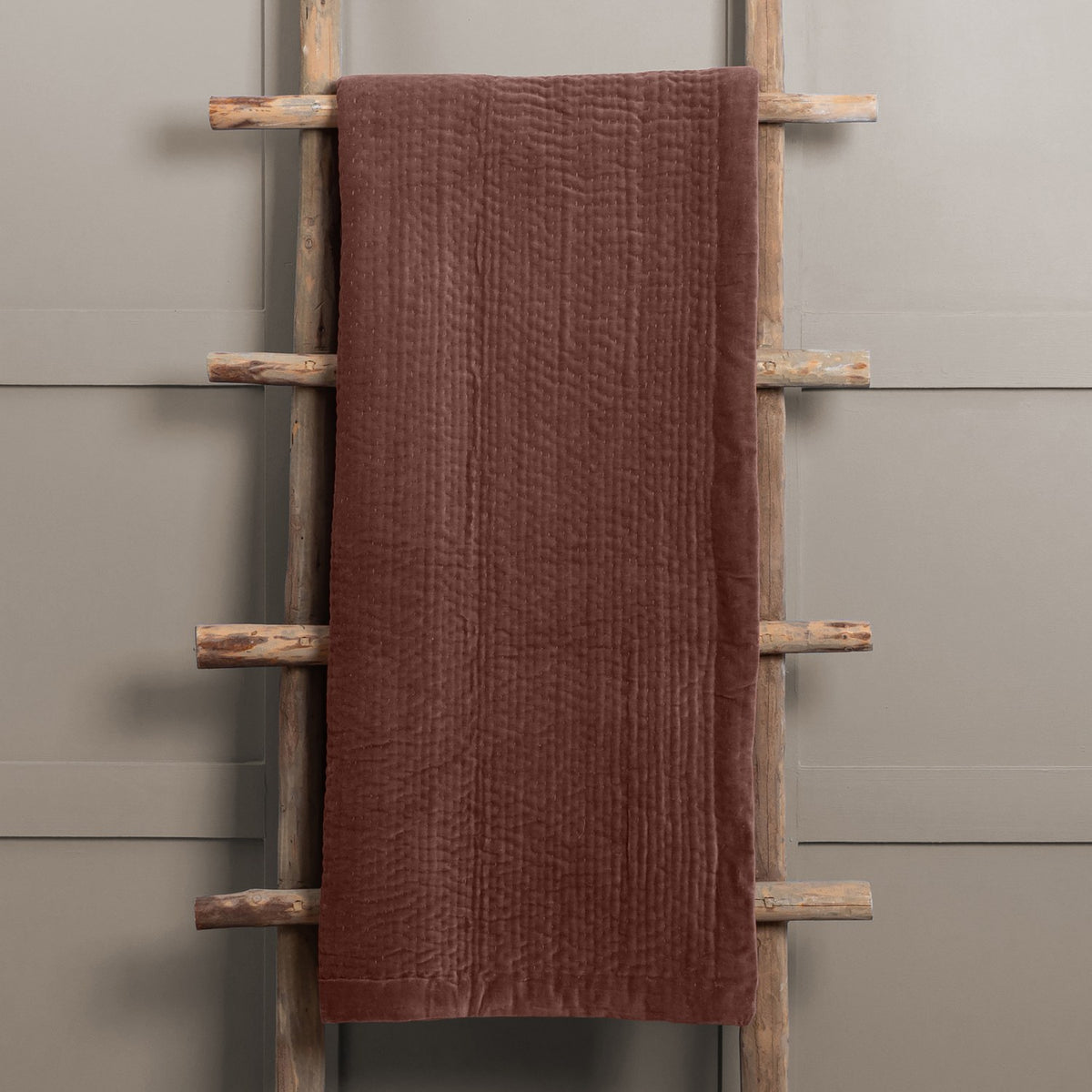 Haze Velvet Cotton Throw in Persimmon, a hand-quilted brown cotton velvet throw with rippled texture, draped over a wooden ladder against a grey wall.