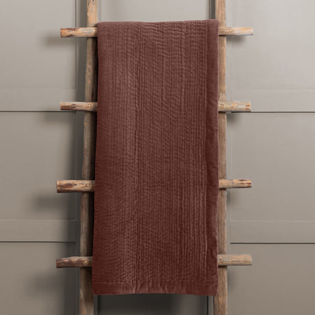 Haze Velvet Cotton Throw in Persimmon, a hand-quilted brown cotton velvet throw with rippled texture, draped over a wooden ladder against a grey wall.