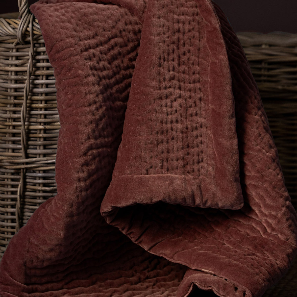 Haze Velvet Cotton Throw in Persimmon, hand-quilted brown-red cotton velvet with rippled texture, draped over a woven wicker basket.