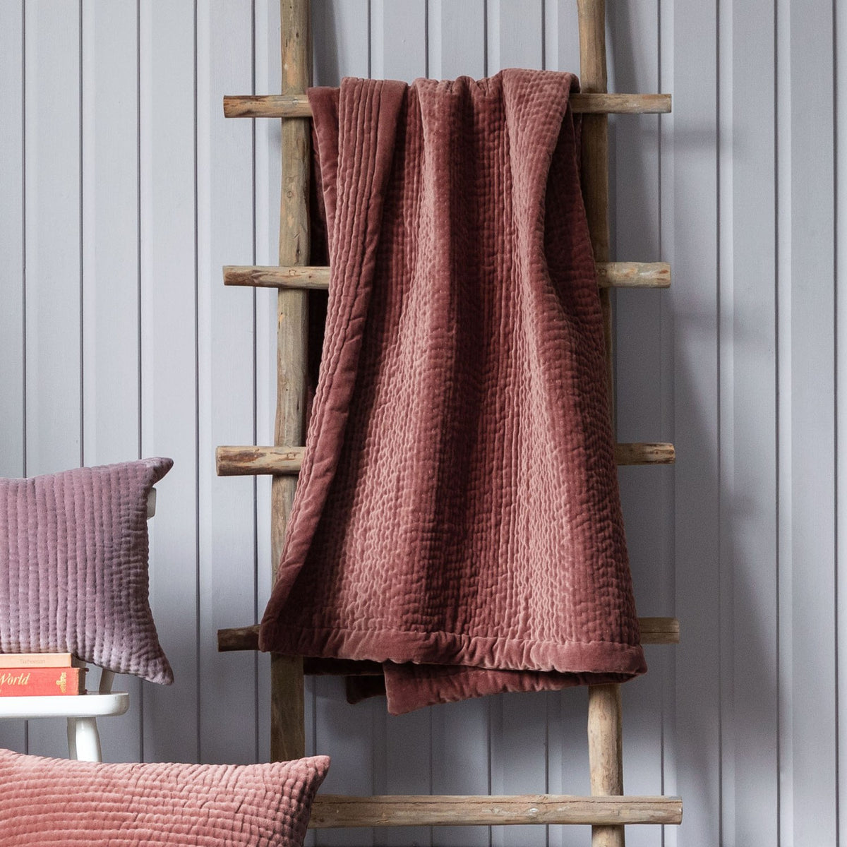 Haze Velvet Cotton Throw in persimmon, hand-quilted with rippled texture, draped over a wooden ladder beside matching cushions against a light grey panelled wall.