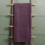 Haze Velvet Cotton Throw in plum, hand-quilted cotton velvet with ribbed texture, draped over a wooden ladder against a green panelled wall.