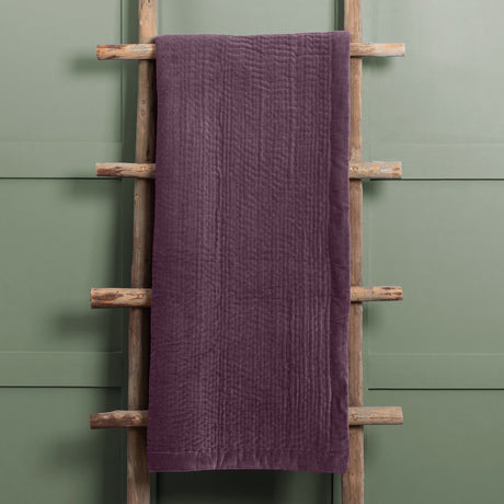 Haze Velvet Cotton Throw in plum, hand-quilted cotton velvet with ribbed texture, draped over a wooden ladder against a green panelled wall.