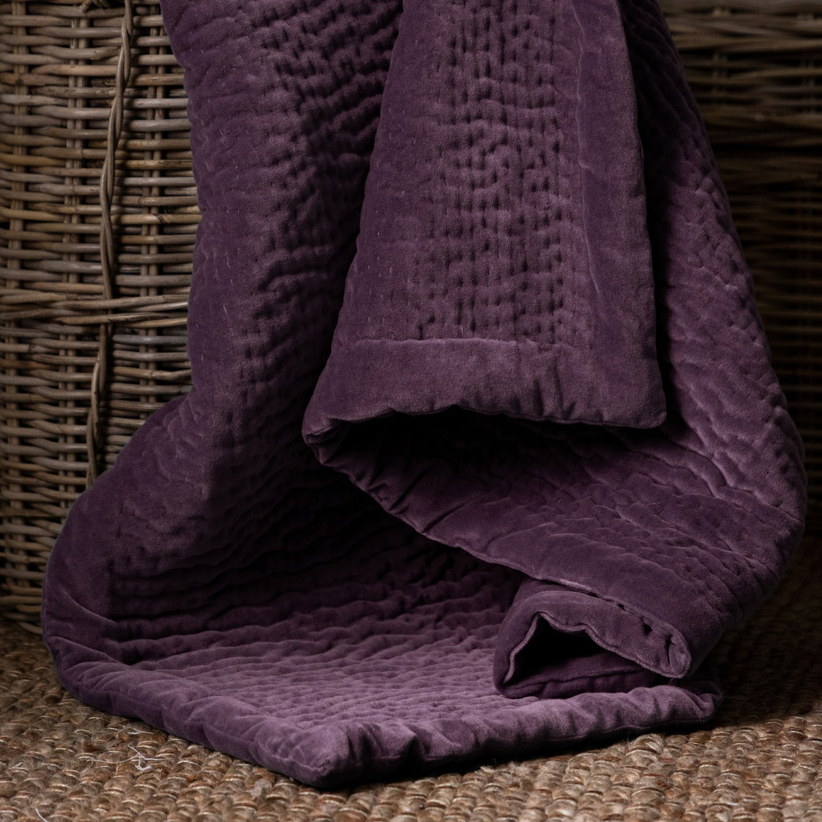 Haze Velvet Cotton Throw in plum, draped over a wicker basket showing hand-quilted rippled texture and folded velvet edge.