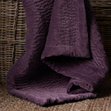 Haze Velvet Cotton Throw in plum, draped over a wicker basket showing hand-quilted rippled texture and folded velvet edge.