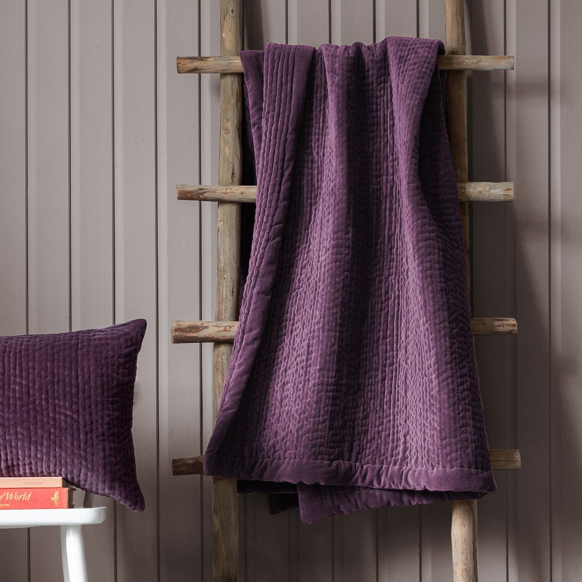 Haze Velvet Cotton Throw in plum draped over a wooden ladder, showing hand-quilted rippled channel texture, with a matching quilted cushion nearby against a grey panelled wall.