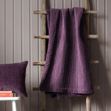 Haze Velvet Cotton Throw in plum draped over a wooden ladder, showing hand-quilted rippled channel texture, with a matching quilted cushion nearby against a grey panelled wall.
