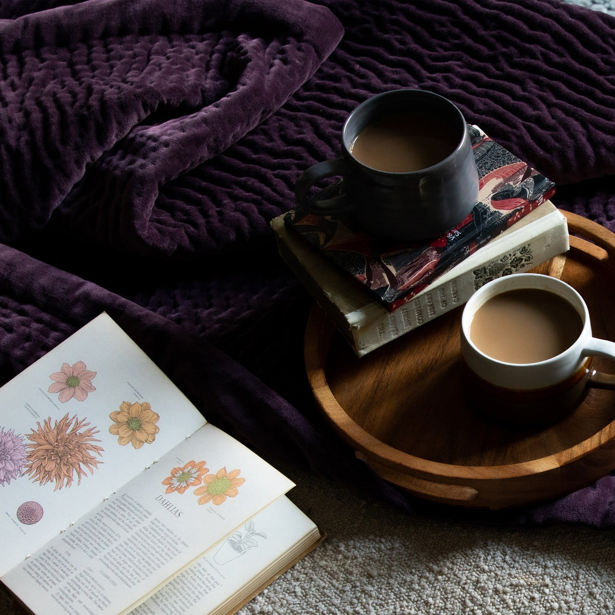 Haze Velvet Cotton Throw in plum, quilted texture draped over a bed, styled with a botanical book and cups of tea on a wooden tray.