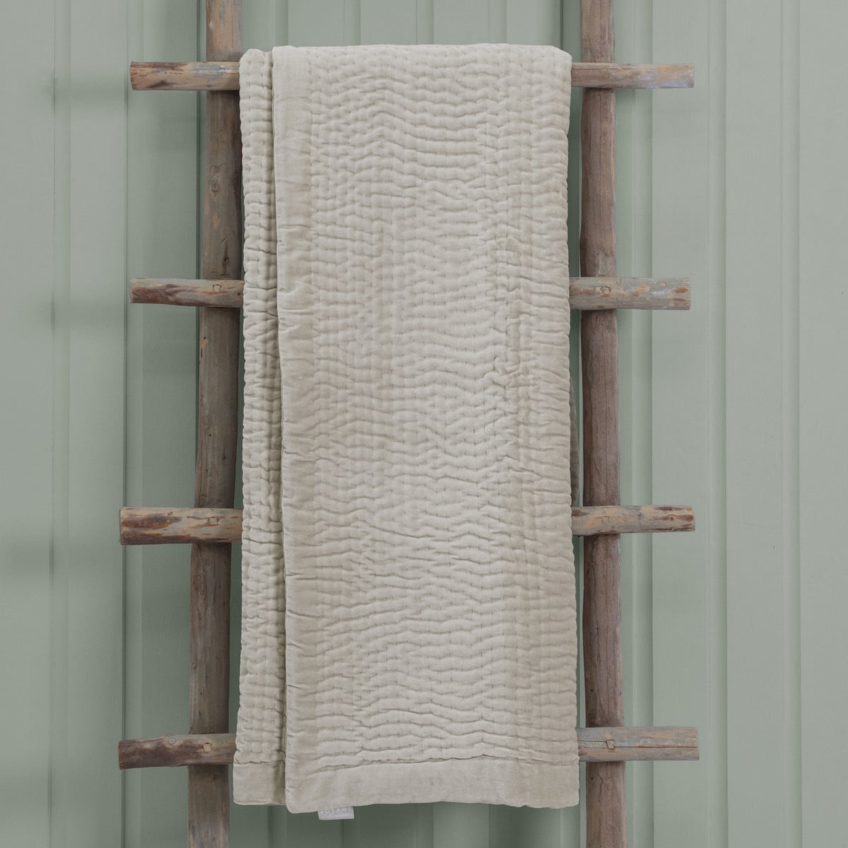 Haze Velvet Cotton Throw in Quartz, a quilted beige cotton throw with wave-like stitched texture, draped over a wooden ladder against a sage green wall.