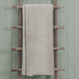 Haze Velvet Cotton Throw in Quartz, a quilted beige cotton throw with wave-like stitched texture, draped over a wooden ladder against a sage green wall.