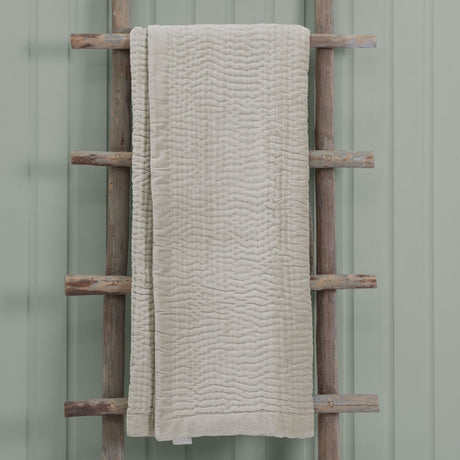 Haze Velvet Cotton Throw in Quartz, a quilted beige cotton throw with wave-like stitched texture, draped over a wooden ladder against a sage green wall.