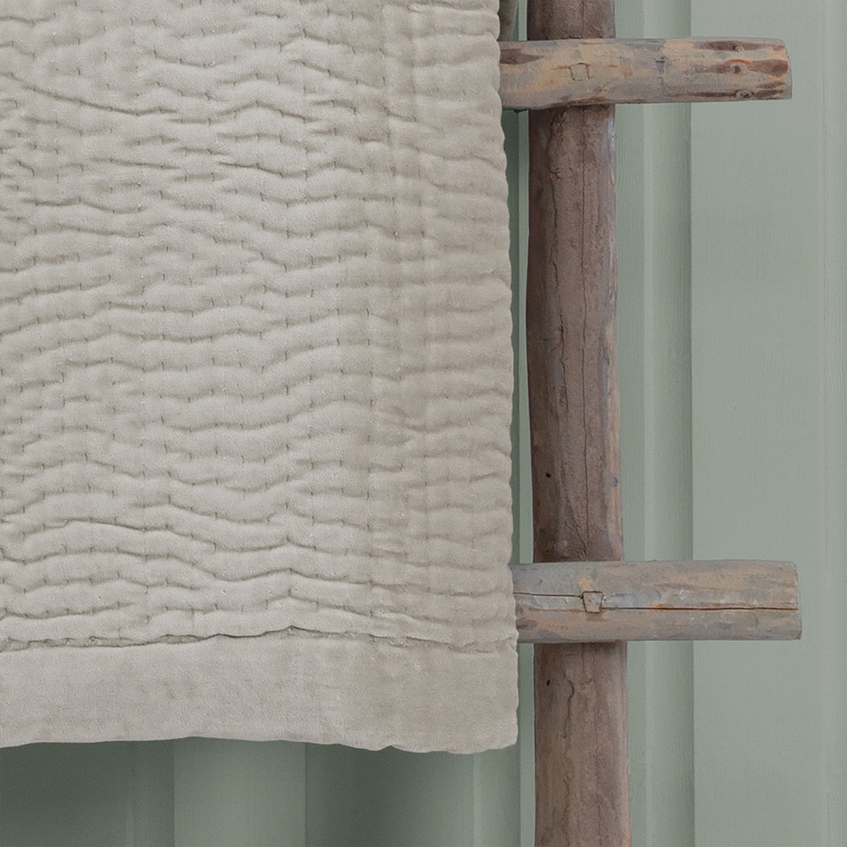 Haze Velvet Cotton Throw in Quartz, a beige quilted cotton throw with wave-like stitched texture, draped over a wooden rack against a sage green wall.