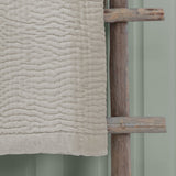 Haze Velvet Cotton Throw in Quartz, a beige quilted cotton throw with wave-like stitched texture, draped over a wooden rack against a sage green wall.