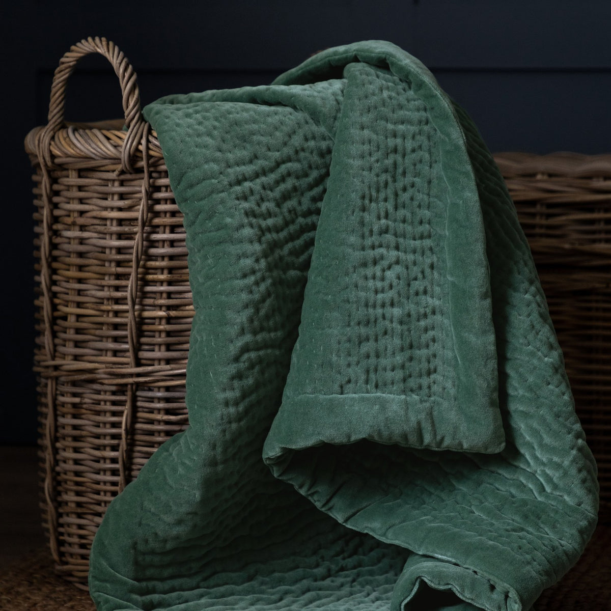 Haze Velvet Cotton Throw in seafoam green, hand-quilted cotton with a soft velvet pile, draped over woven wicker baskets against a dark background.