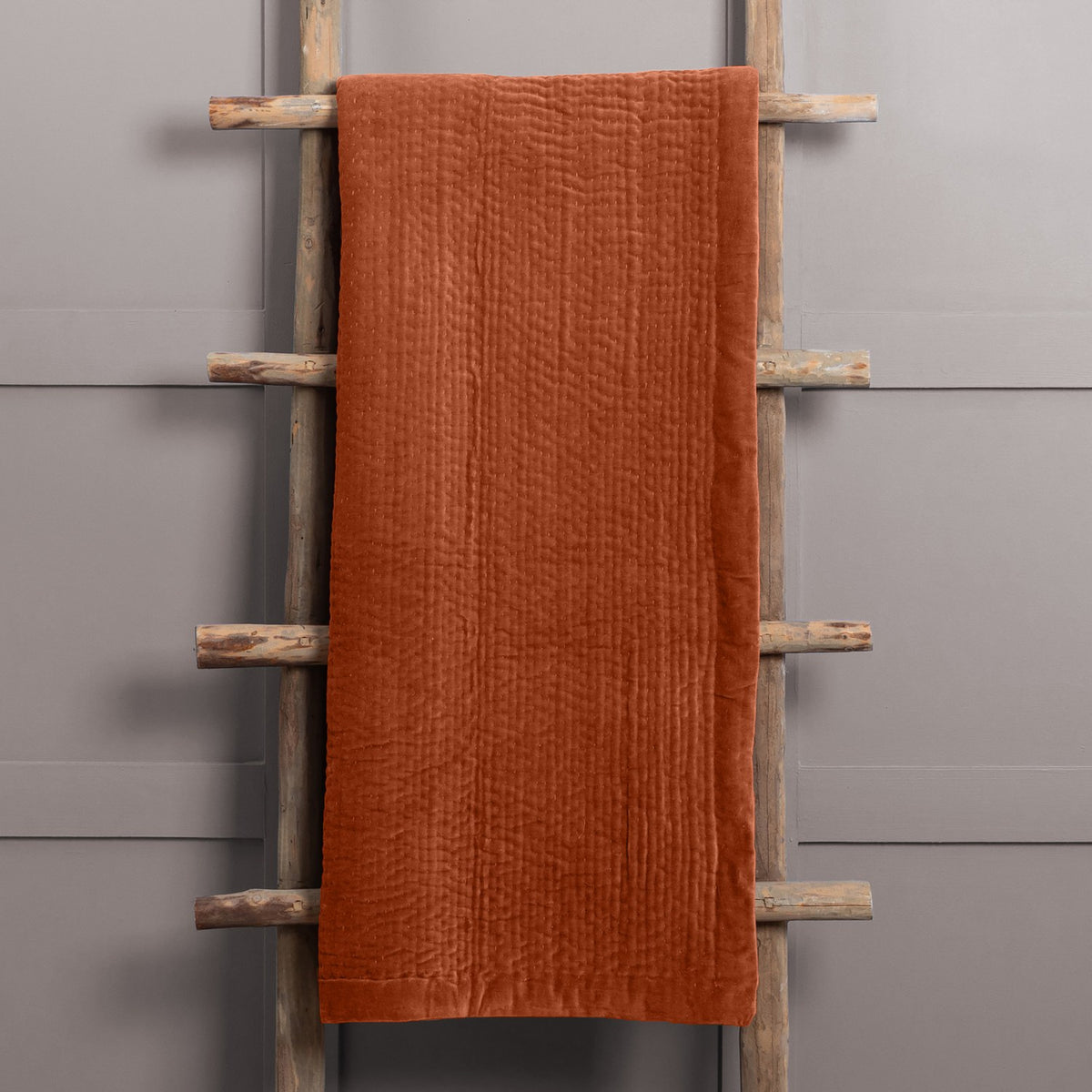Haze Velvet Cotton Throw in Sunset, a rust-orange quilted cotton velvet throw with rippled channel texture, draped over a wooden ladder against a grey background.