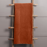 Haze Velvet Cotton Throw in Sunset, a rust-orange quilted cotton velvet throw with rippled channel texture, draped over a wooden ladder against a grey background.